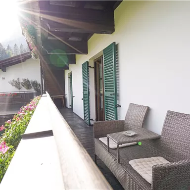A sunny balcony with modern rattan furniture and colorful flowers. The shutters are green and the surroundings are rural and beautiful.