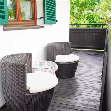 A cozy balcony with two rattan chairs and a small table. In the background, there are green plants and windows with wooden shutters.