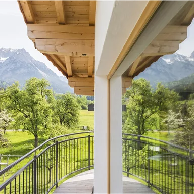 Eine moderne Balkonansicht mit Blick auf grüne Wiesen und majestätische Berge. Die sanfte Natur ist von Bäumen umgeben und der Himmel ist klar.