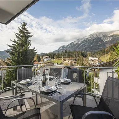 A cozy balcony with a table and chairs, perfect for outdoor meals. In the background, majestic mountains and green trees can be seen.