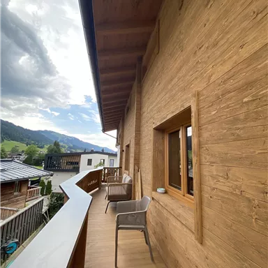 Ein großzügiger Balkon mit Holzverkleidung und einem Blick auf die Berge. Im Hintergrund sind weitere Häuser und ein bewölter Himmel zu sehen.