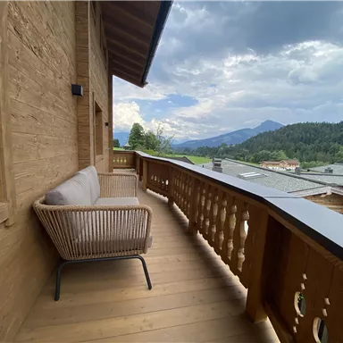 Ein Balkon aus Holz mit einem gemütlichen Sessel und Blick auf grüne Hügel und Berge. Der Himmel ist leicht bewölkt und bietet eine entspannte Atmosphäre.