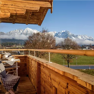 A balcony with wooden cladding and two chairs. In the background, snow-covered mountains and a clear sky can be seen.