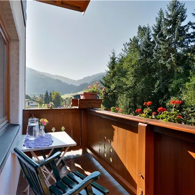 Ein gemütlicher Balkon mit einem Tisch und Stühlen. Der Blick reicht auf grüne Bäume und Berge im Hintergrund.