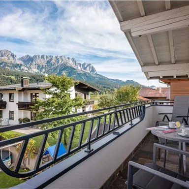 A cozy balcony view with chairs and a table. Impressive mountains and green trees are visible in the background.