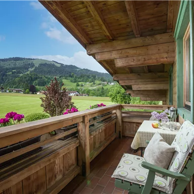Ein gemütlicher Balkon mit Blick auf grüne Wiesen und Berge. Die Blumen und das rustikale Holz schaffen eine einladende Atmosphäre.