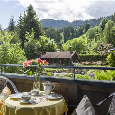A cozy balcony with a covered table and a view of green mountains. On the table, there is a flower vase, cups, and dishes.