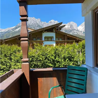 View from a balcony onto a green hedge and impressive mountains in the background. The sun is shining under a bright blue sky.
