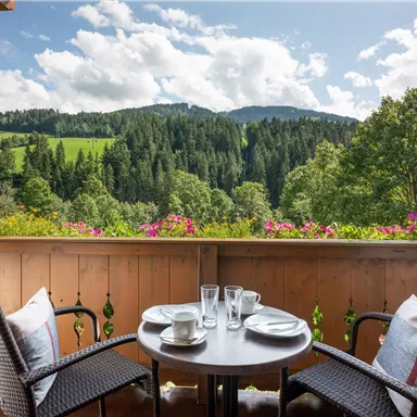 A balcony with a small table and two chairs. In the background, green hills and forests are visible under a bright sky.