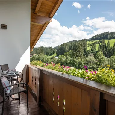 A beautiful balcony with two chairs and a table, surrounded by colorful flowers. In the background, a green landscape stretches under a clear sky.