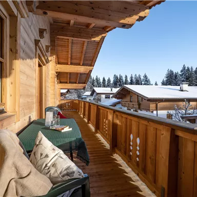A wooden balcony with a table and chairs. The surroundings are covered in snow and surrounded by trees.