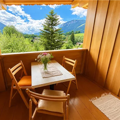 A cozy balcony with wooden paneling and a view of green mountains and trees. On the table, there are flowers in a vase and two chairs are grouped around the table.