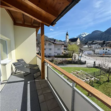 A balcony with loungers and a view of a picturesque village in the mountains. The surroundings are green and the sky is clear.