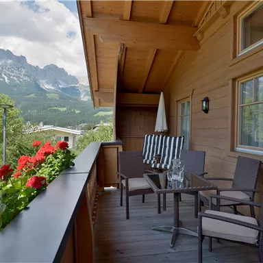 A cozy balcony with chairs and a table, surrounded by blooming geraniums. In the background, majestic mountains and a blue sky are visible.
