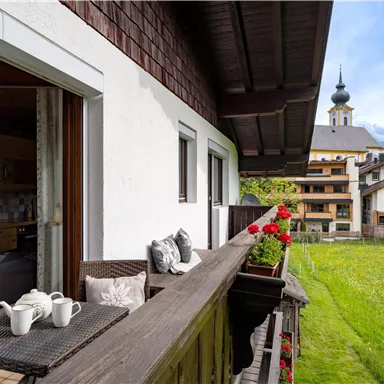 Ein Balkon mit gemütlichen Möbeln und blühenden Blumen. Im Hintergrund sind malerische Gebäude und eine Kirche zu sehen.