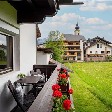 Ein Balkon mit Blick auf eine grüne Wiese und ein paar Häuser im Hintergrund. Rote Blumen zieren das Geländer, und der Himmel ist teilweise bewölkt.
