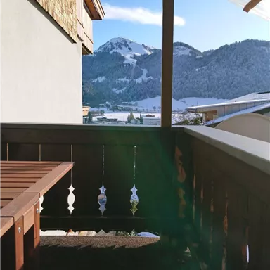 A cozy balcony with a wooden railing and table. In the background, snow-covered mountains and a blue sky can be seen.