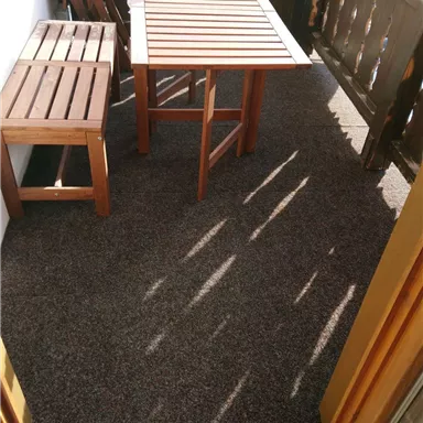 A small balcony with a table and two chairs. The floor is covered with a carpet, and sunshine can be seen.