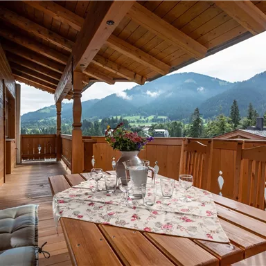 Ein gemütlicher Balkon mit Holzverkleidung und Blick auf grüne Berge. Auf dem Tisch steht eine Blumenvase und mehrere Gläser.