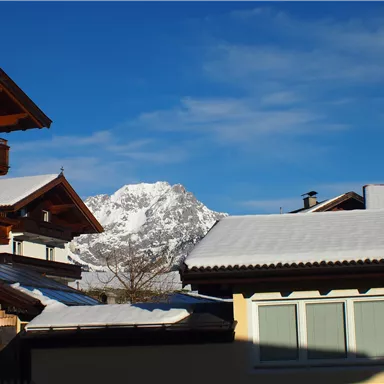 A winter landscape with snow-covered roofs and mountains in the background. The sky is clear and blue.