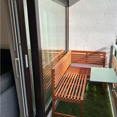 A small balcony with a wooden bench and a table. The floor is covered with green carpet and large windows let in a lot of light.