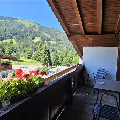 A balcony with a table and two chairs, surrounded by colorful flowers. In the background, green mountains and a clear sky can be seen.