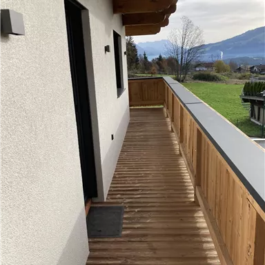 A wooden balcony with a view of a green meadow and mountains in the background. The wall is light and the sky is partly cloudy.