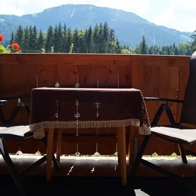 A cozy balcony with two chairs and a small table. In the background, a beautiful mountain landscape and trees can be seen.