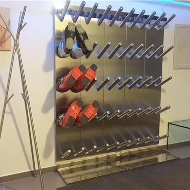 A modern shoe rack made of metal with several bars for hanging shoes. Red and black shoes are visible, as well as a minimalist coat rack and a glass table in the room.