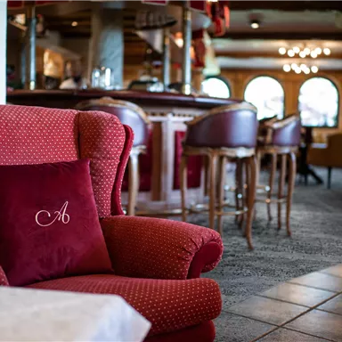 Ein gemütlicher Lounge-Bereich mit einem roten Sessel und einem Kissen mit dem Buchstaben "A". Im Hintergrund sind Barhocker und eine einladende Atmosphäre sichtbar.