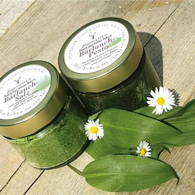 Two jars of wild garlic pesto are sitting on a wooden table. Fresh wild garlic leaves and white daisies are arranged around the jars.