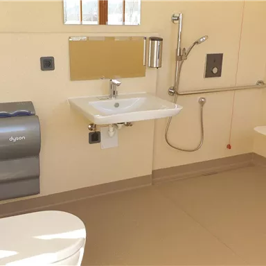 Ein modernes Badezimmer mit einer Toilette, einem Waschbecken und Haltegriffen. Die Wände sind hell und der Raum ist gut beleuchtet.