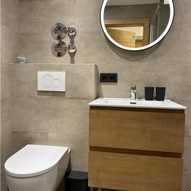 A modern bathroom with a wooden vanity, a sink, and a round mirror. The walls are clad with gray tiles, and there is a simple toilet.
