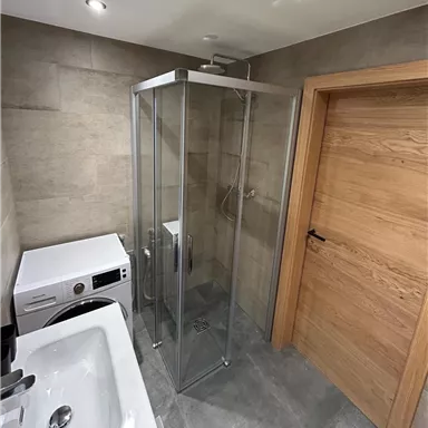 A modern bathroom with a glass shower enclosure and a washing machine. The walls are covered with gray tiles and the door is made of wood.