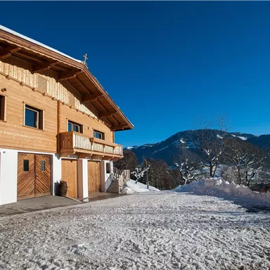 Ein traditionelles Holzhaus in der winterlichen Landschaft. Im Hintergrund sind schneebedeckte Berge und ein klarer blauer Himmel zu sehen.