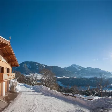 Ein Chalet in den Bergen, umgeben von Schnee und Bäumen. Der klare Himmel und die sonnige Aussicht schaffen eine schöne, winterliche Atmosphäre.