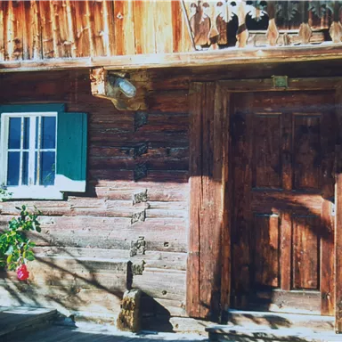 Ein rustikales Blockhaus mit einer Holztür und einem kleinen Fenster. Rosenblüten ranken sich an der Wand.