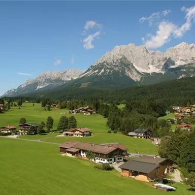 Eine malerische Landschaft mit grünen Wiesen und verstreuten Häusern. Im Hintergrund erheben sich beeindruckende Berge unter einem klaren Himmel.