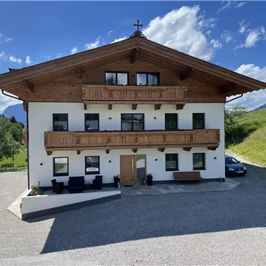 A picturesque building in alpine style with wooden balustrades and green lawn. The surroundings are filled with mountains and a radiant blue sky.
