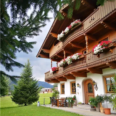 A traditional chalet with blooming balconies and a beautiful garden. Gentle hills can be seen in the background.