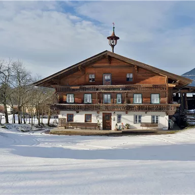 Ein traditionelles Holzhaus im Schnee, umgeben von schneebedeckter Landschaft. Im Hintergrund sind Berge und ein teilweise bewölkter Himmel zu sehen.