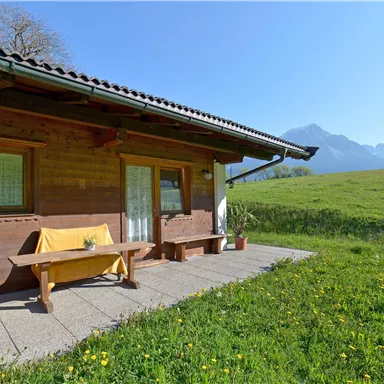 Ein gemütliches Holzhaus mit einer Terrasse, umgeben von grünen Wiesen und Bergen im Hintergrund. Der Himmel ist klar und sonnig.