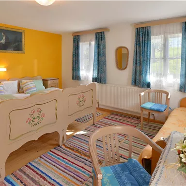 Ein gemütliches Schlafzimmer mit einem Doppelbett und bunten Kissen. Die Wände sind in warmen Farben gehalten und große Fenster sorgen für viel Licht.