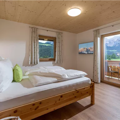 A cozy bedroom with a wooden bed and white bedding. The windows offer a view of the mountains and a small balcony.