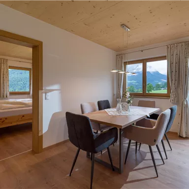 A modern dining room with a large table and stylish chairs. Through the windows, there is a beautiful view of the mountains and the green landscape visible.
