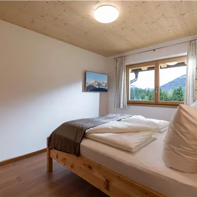 A cozy bedroom with a large window and a view of the mountains. The room is bright and simply furnished with a wooden bed and fresh linens.