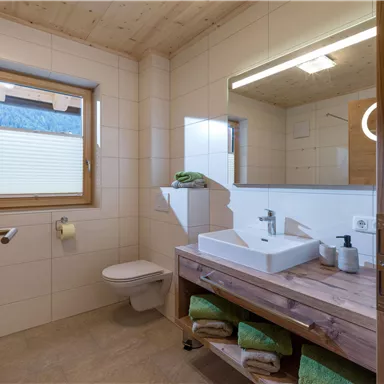 A modern bathroom with wood and tile accents. There is a large mirror surface, a sink, and fresh towels in green.