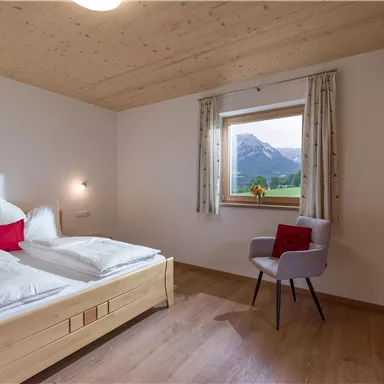 A cozy bedroom with a double bed and a chair. Through the window, you can see a beautiful mountain landscape.