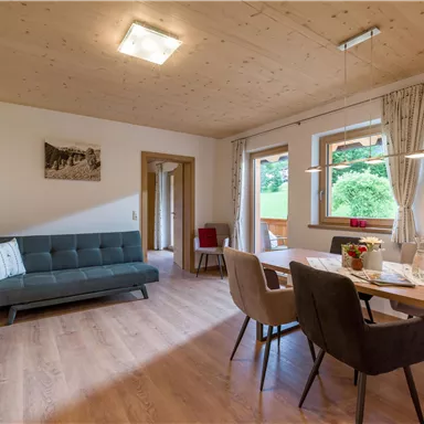 A modern living room with a sofa and a dining table. Large windows let in plenty of light and provide a view of the green surroundings.
