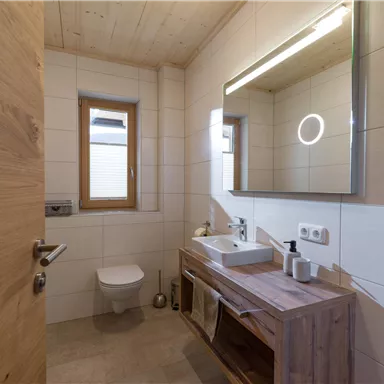 A modern bathroom with wood and tile elements. It features a sink, a mirror with lighting, and a window for natural light.